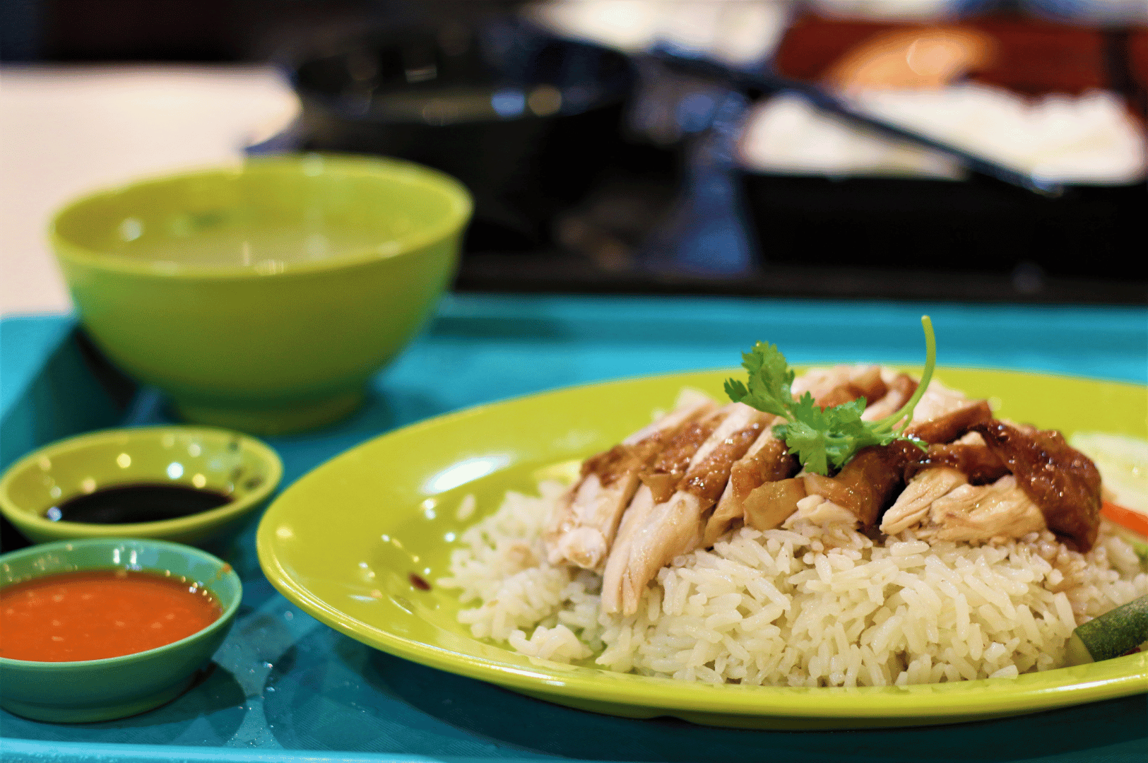 Classic serving of Hainanese chicken rice on a green plate with brown sauce and cilantro garnish, accompanied by chili, soy, and soup in matching bowls—presented on a blue tray at Maxwell Food Center, showcasing the dish’s balanced flavors and iconic simplicity.