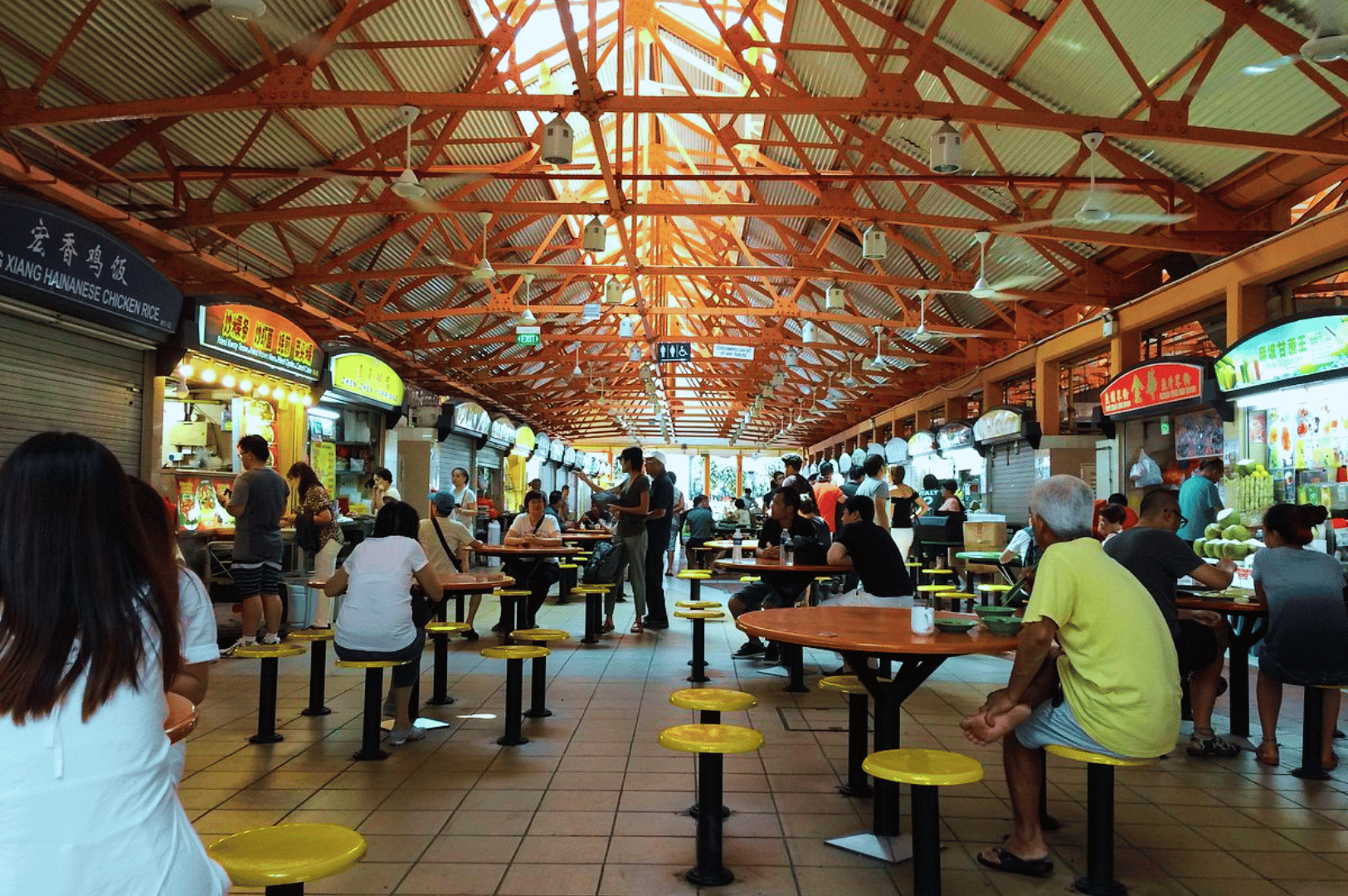 Wide-angle view of Maxwell Food Center’s open-air hawker hall, framed by orange metal trusses and ceiling fans—diners gather around yellow stools and round tables, surrounded by brightly lit food stalls offering diverse Southeast Asian dishes in a lively, communal setting.