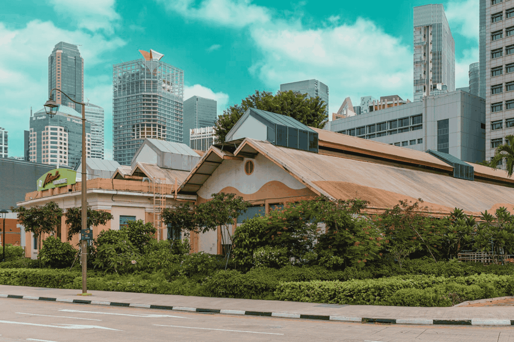 Juxtaposition of heritage and modernity near Maxwell Food Center—traditional sloped-roof building nestled in greenery stands before a skyline of glass-clad skyscrapers, reflecting Singapore’s fusion of cultural preservation and urban innovation under a bright, cloud-speckled sky.