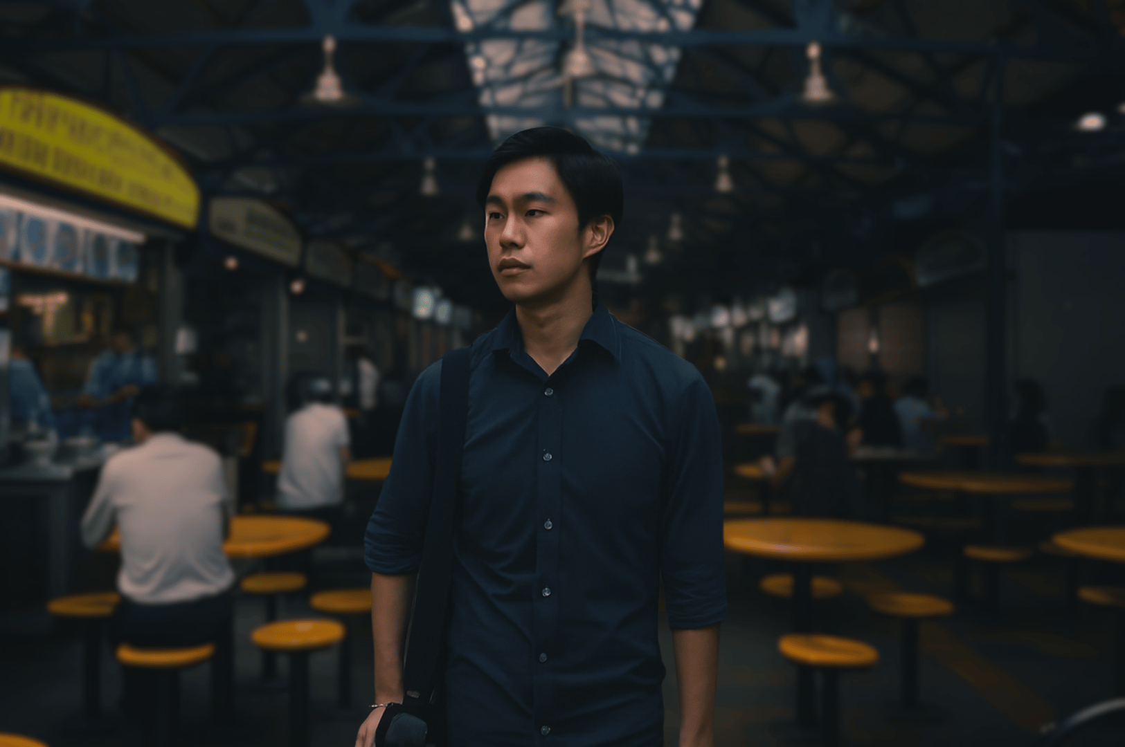 Young diner standing amid yellow tables and stools inside Maxwell Food Center’s bustling hawker hall, framed by metal beams and hanging lights—capturing the casual, communal spirit of Singapore’s street food culture.
