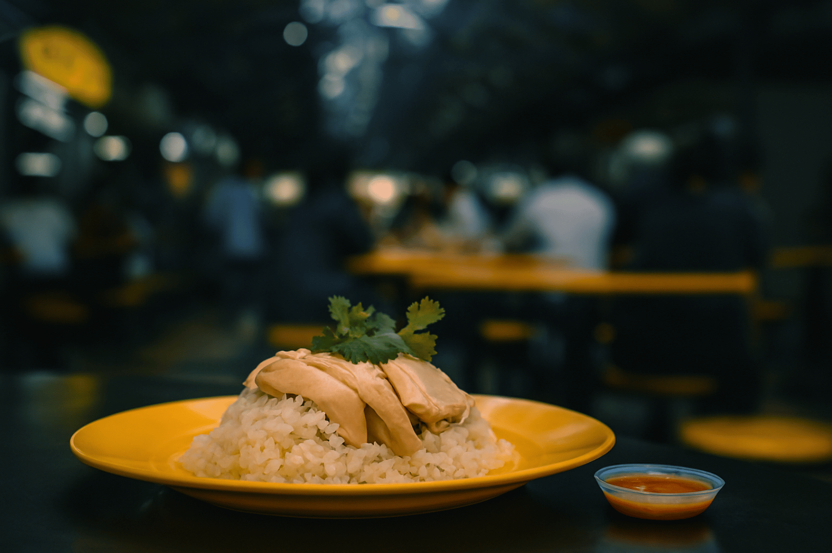 Close-up of Hainanese chicken rice served on a vibrant yellow plate at Maxwell Food Center—poached chicken over fragrant rice, garnished with cilantro and paired with chili sauce, set against the blurred bustle of communal hawker dining.