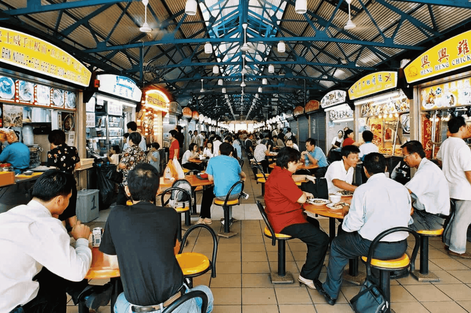 Wide-angle view of Maxwell Food Center’s bustling indoor hawker hall, framed by a blue steel ceiling and lined with yellow-signed food stalls serving Chinese specialties—office workers and locals gather at tiled tables, reflecting Singapore’s vibrant midday dining culture.