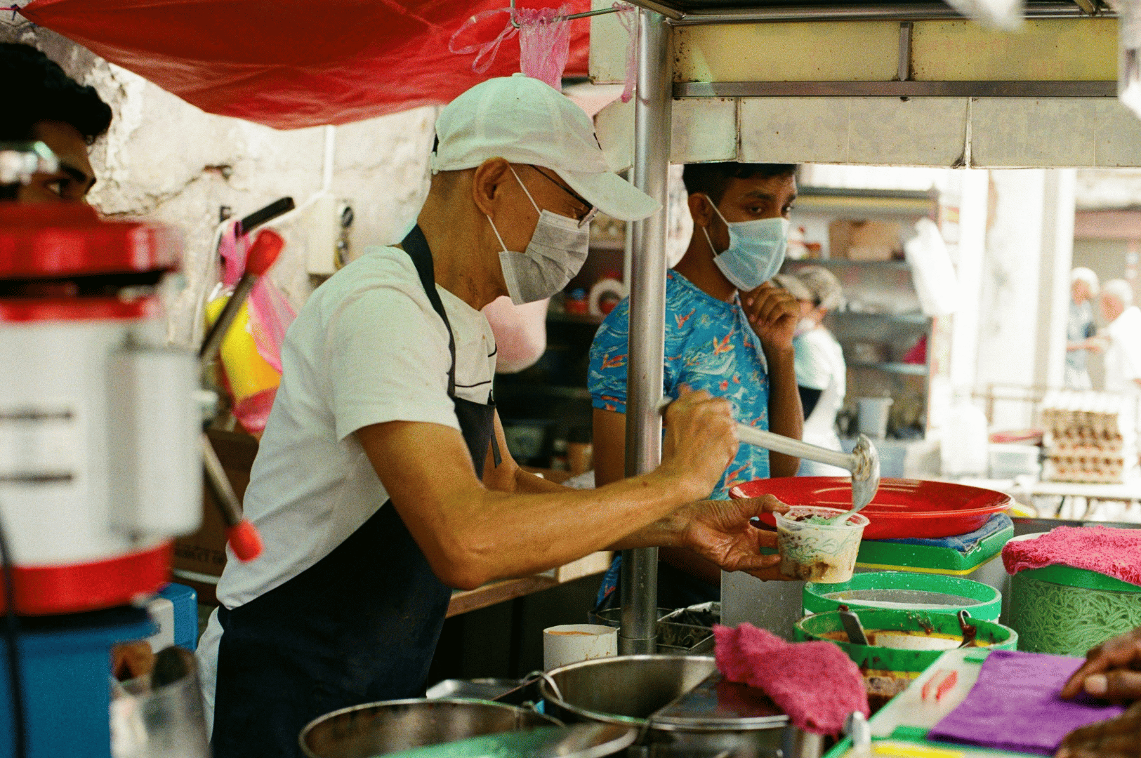 Street food vendor in white cap and apron ladling hot dishes at Maxwell Food Center, surrounded by colorful towels and cooking tools—capturing the hands-on craft and everyday rhythm of Singapore’s hawker heritage.