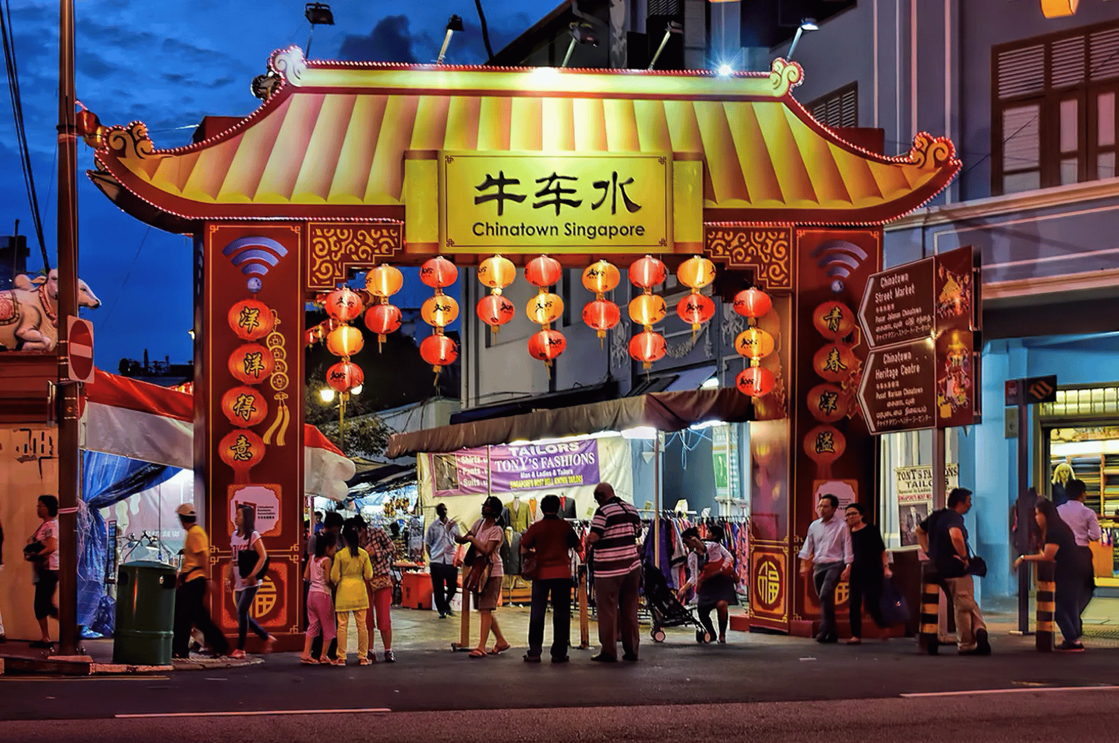 Evening view of Chinatown Singapore’s ornate entrance archway, glowing with red lanterns and golden roof tiles—families and tourists stroll beneath colonial facades and festive lights, capturing the vibrant cultural heartbeat just steps from Maxwell Food Center’s hawker treasures.