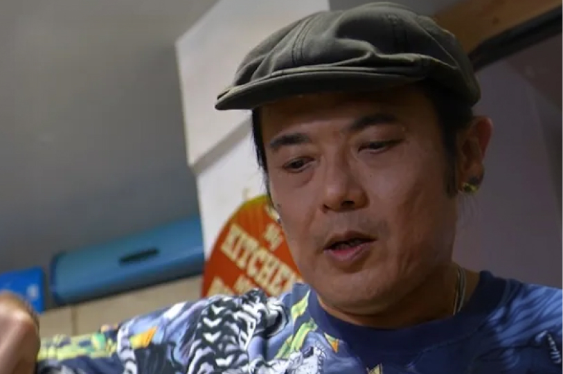 A close-up portrait of Chef Chris Choo wearing a dark olive flat cap and a patterned blue shirt, looking down as he prepares a meal.