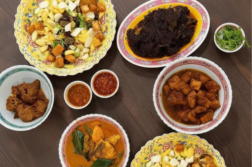 An overhead view of a Peranakan spread on a wooden table, featuring fruit rojak, beef rendang, curry fish, and various spicy sambals in traditional ornate bowls.