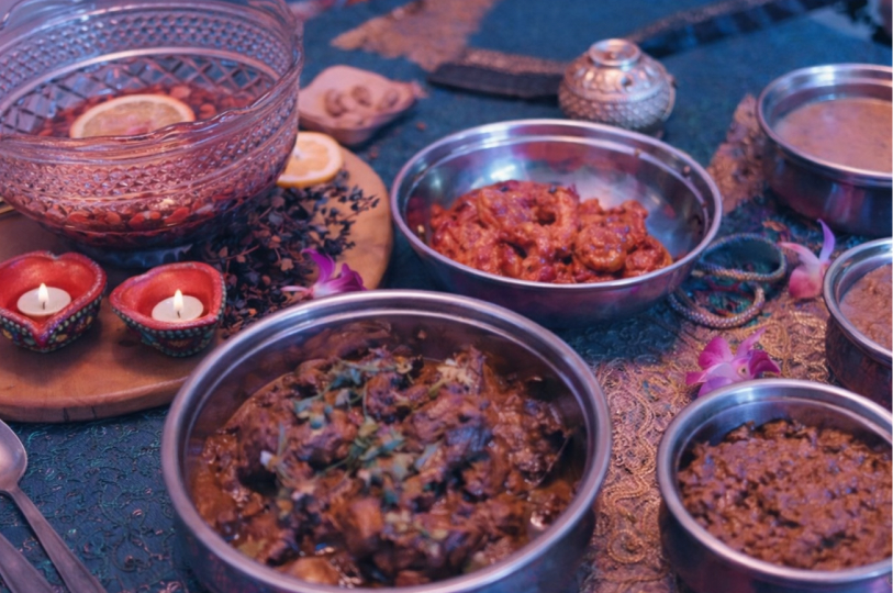 A close-up of several metal bowls containing different braised meats and stews, set on a blue patterned tablecloth with flickering tea light candles in the background.