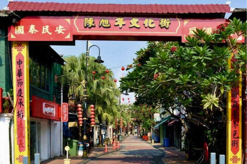 A vibrant red and yellow Chinese ceremonial archway framing a tree-lined street with hanging red lanterns.