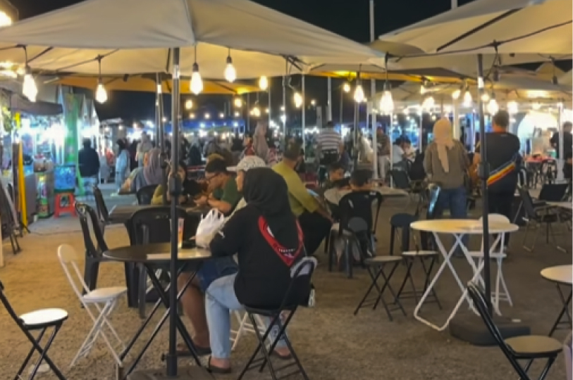A crowded outdoor night market with people sitting at tables under large white umbrellas and warm hanging lights.