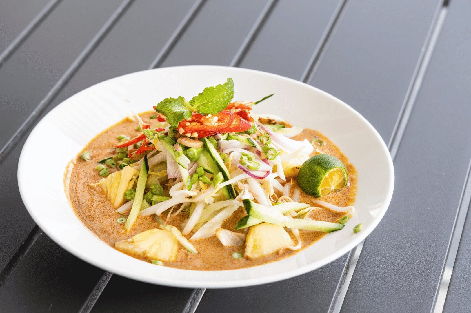 Overhead view of a vibrant bowl of Johor-style laksa, set against a dark gray slatted surface. The rich reddish-brown broth cradles fresh bean sprouts, cucumber, pineapple, red chilies, and mint, with a halved calamansi lime poised for a citrusy kick. Garnished with peanuts and green onions, the dish bursts with color and texture—capturing the bold, tropical flavors that define Johor Bahru’s street food scene.
