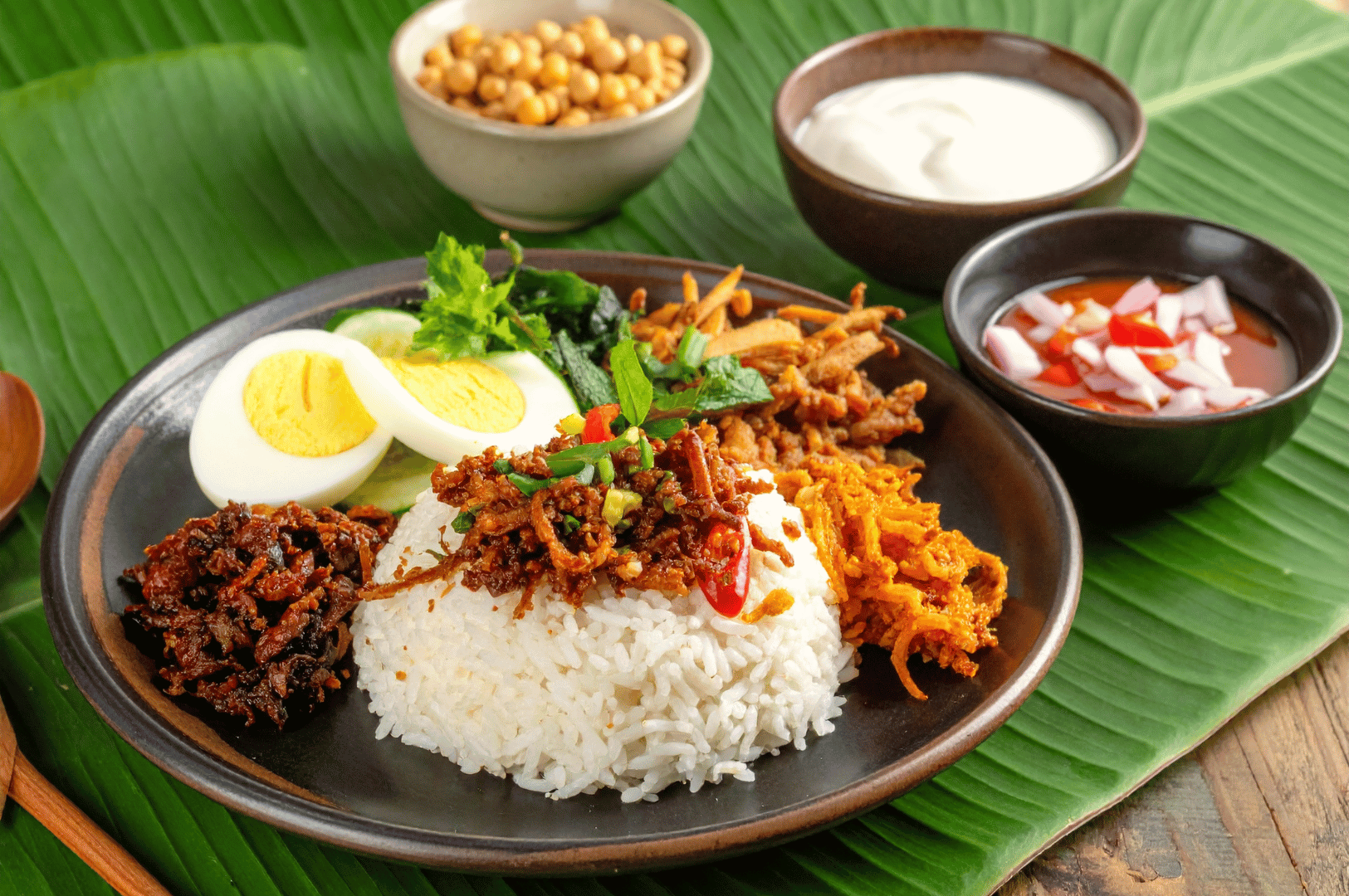 Traditional Nasi Lemak artfully plated on a banana leaf-lined dark dish. A fragrant mound of white rice anchors the composition, encircled by sambal, crispy anchovies, roasted peanuts, cucumber slices, and hard-boiled egg. A portion of spiced meat—possibly rendang—adds richness, while small bowls of coconut cream and chili-onion sambal in the background hint at customizable flavor. This iconic Malaysian breakfast captures the soulful balance of heat, texture, and heritage central to Johor Bahru’s culinary identity.