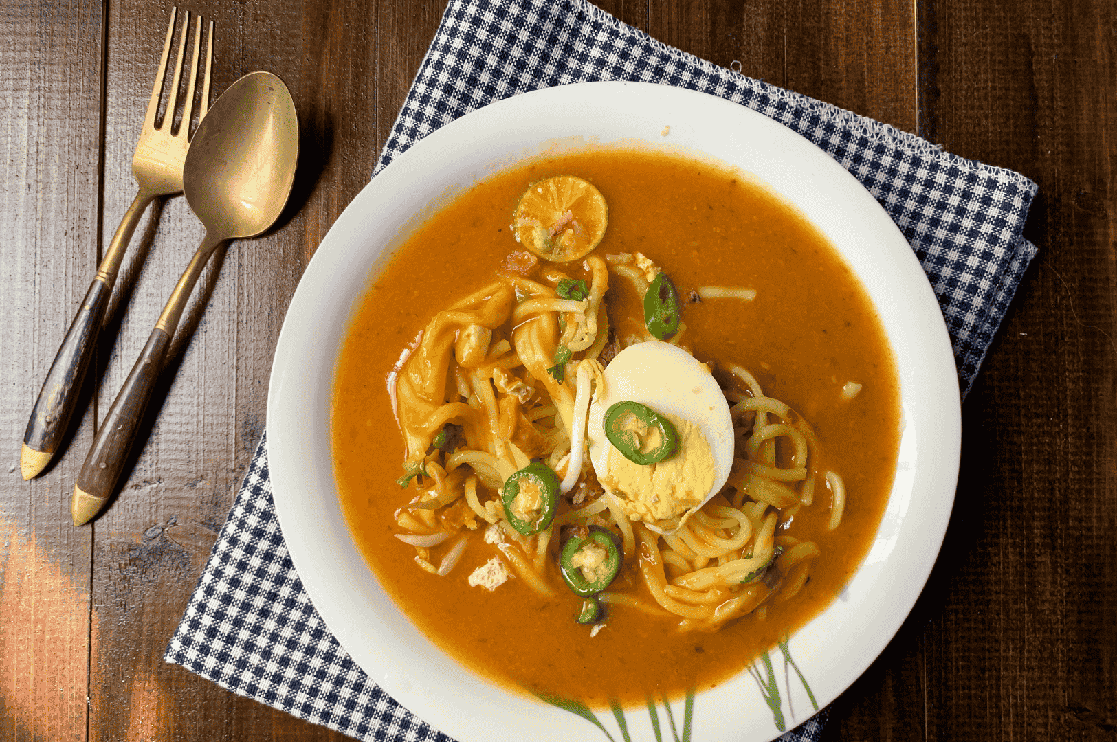 Bowl of thick yellow noodles in a rich, curry-like broth, garnished with green chili slices, a halved boiled egg, lime wedge, and shredded meat. Presented on a white plate over a blue checkered cloth, the dish evokes warmth and comfort. The golden spoon and fork beside it hint at a homey yet elevated dining experience—capturing the soulful spice and layered flavors of Johor Bahru’s culinary heritage.