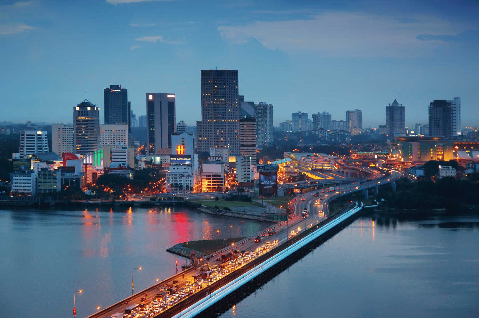Twilight cityscape of Johor Bahru, with a brightly lit causeway leading into a bustling skyline of high-rise buildings and active traffic. Reflections shimmer across the water, amplifying the energy of the evening rush. This luminous urban scene marks the close of a flavorful day trip—where culinary exploration meets the pulse of a city in motion.