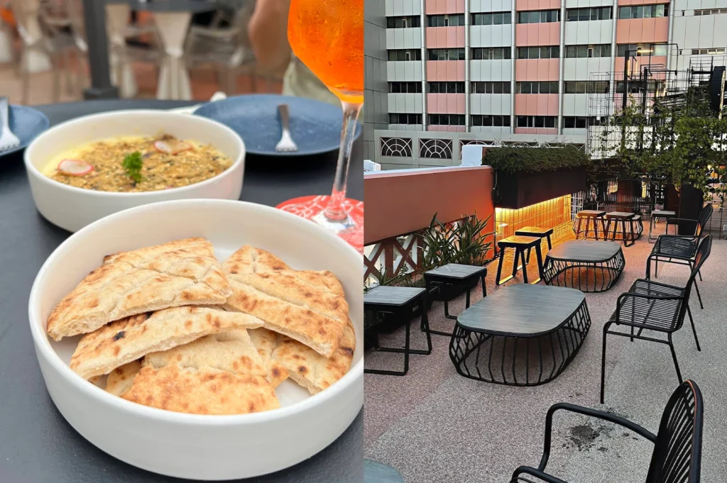 The left side of this image shows a close-up of a white bowl filled with warm, toasted pita bread triangles, with a secondary bowl of creamy dip topped with radish and herbs visible in the background. A glass of vibrant orange Aperol Spritz sits partially in frame. The right side depicts an empty, modern rooftop terrace during the daytime. The area is furnished with black wire-frame chairs and low, dark coffee tables arranged on a speckled grey and pink floor. A tall building with a pink and white facade stands directly behind the terrace, adding a geometric, urban feel to the space.