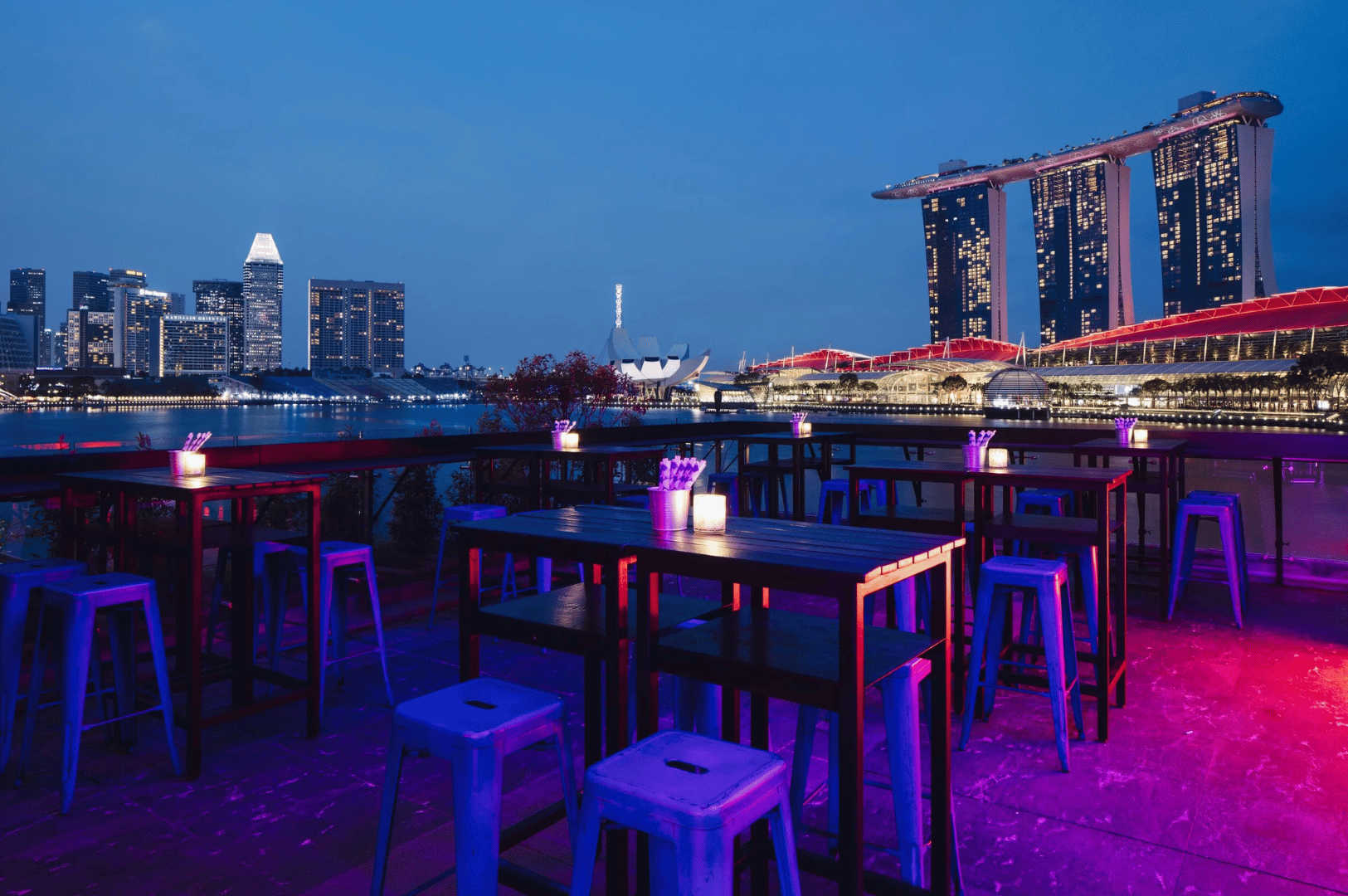 Rooftop bar with modern tables and chairs overlooking Singapore’s Marina Bay skyline at dusk, featuring illuminated skyscrapers and calm waterfront views.