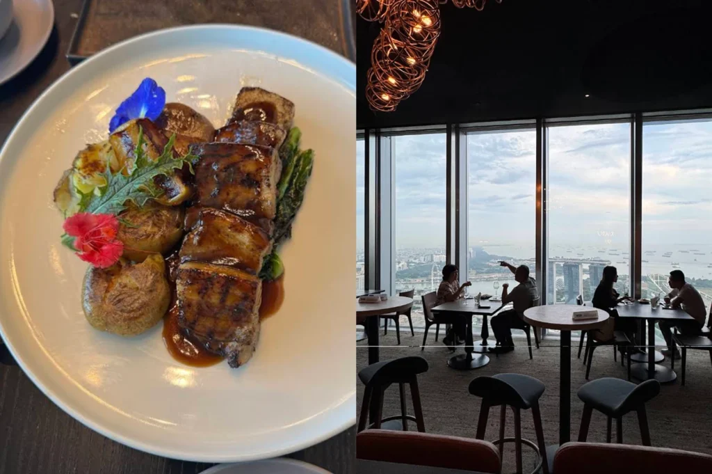 Formal indoor dining aesthetic. On the left, a plate features thick, square-cut pieces of glazed pork belly or braised meat, garnished with a bright blue butterfly pea flower and a red hibiscus. It is served with roasted potatoes and leafy greens. On the right, the interior of a high-rise restaurant is shown, featuring floor-to-ceiling windows that offer an expansive view of a bay filled with ships and the iconic Marina Bay Sands hotel. Diners are silhouetted against the daylight, seated at round wooden tables beneath unique, spiraling copper light fixtures.