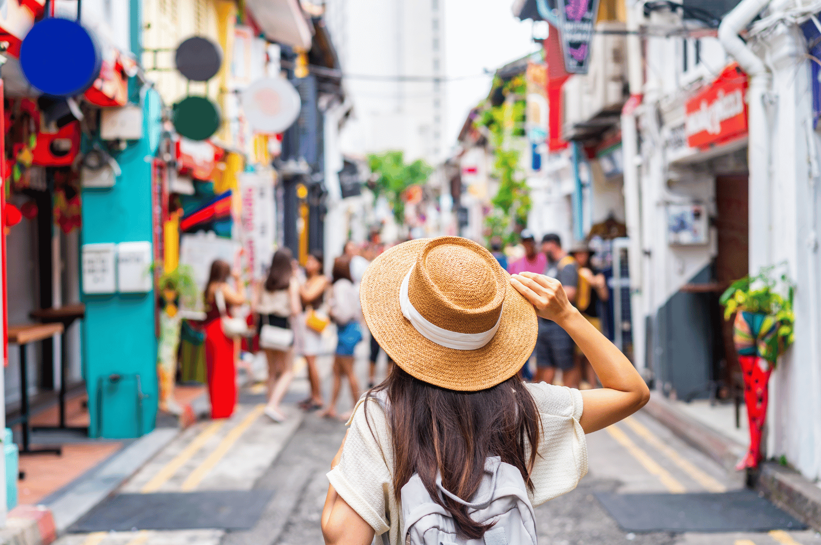 Daytime street scene in one of Singapore’s alternative shopping districts, featuring a traveler in a straw hat and backpack exploring a vibrant row of heritage shophouses and indie boutiques. The mix of traditional architecture, colorful facades, and local signage evokes a slower, more culturally immersive retail experience beyond Orchard Road’s glitz.