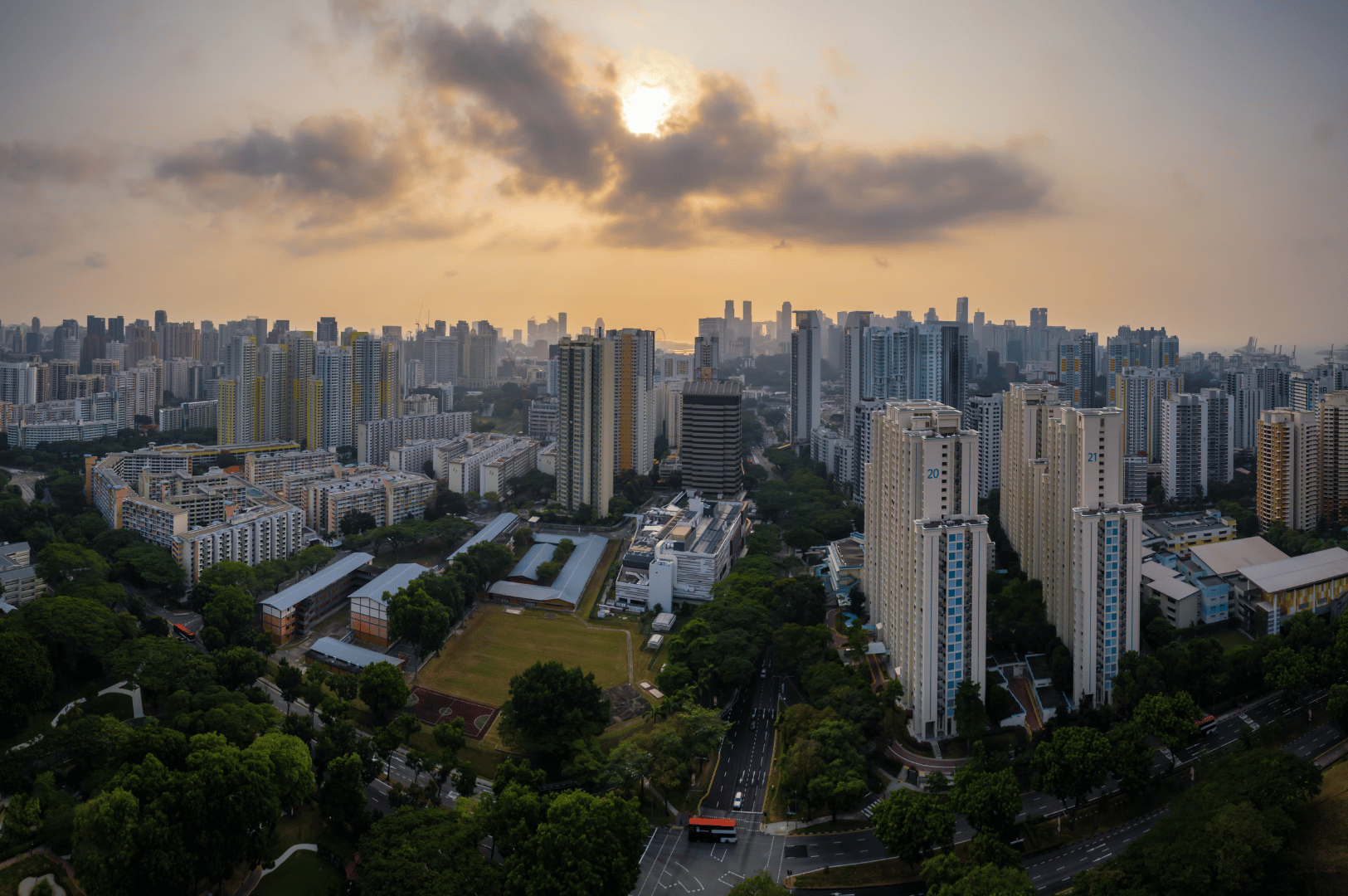 Panoramic sunset view of a residential district in Singapore, with high-rise buildings bathed in golden light and a red bus winding through tree-lined streets. This quieter, lived-in urban landscape offers a glimpse into the everyday rhythms and local shopping enclaves beyond Orchard Road’s commercial glamour.