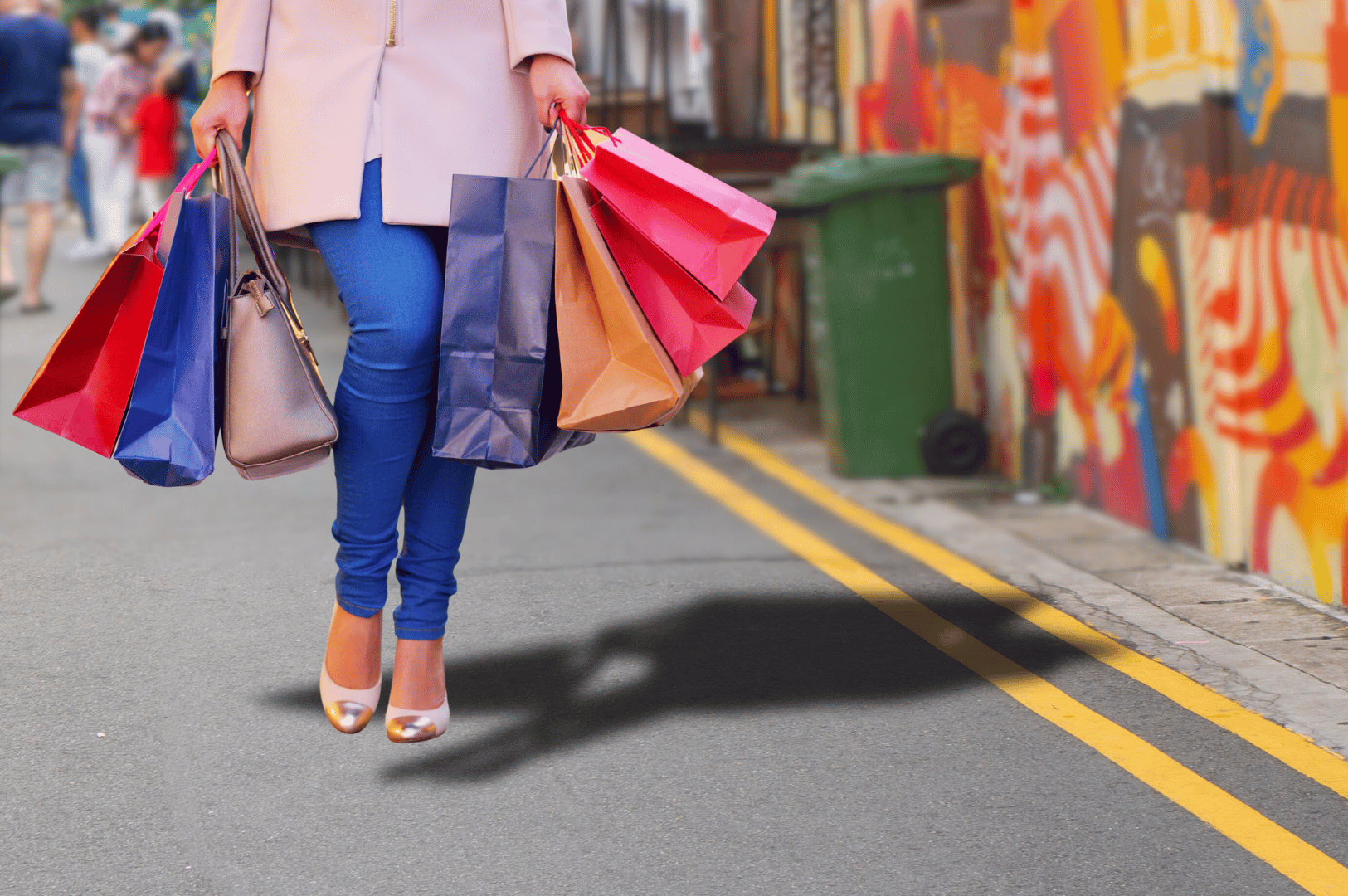 Stylish shopper walking down a mural-lined street in Singapore, carrying a cascade of colorful shopping bags in both hands. Dressed in a light coat and metallic-tipped heels, the figure evokes the fashion-forward spirit of local retail enclaves beyond Orchard Road, where street art and boutique finds blend into vibrant urban life.
