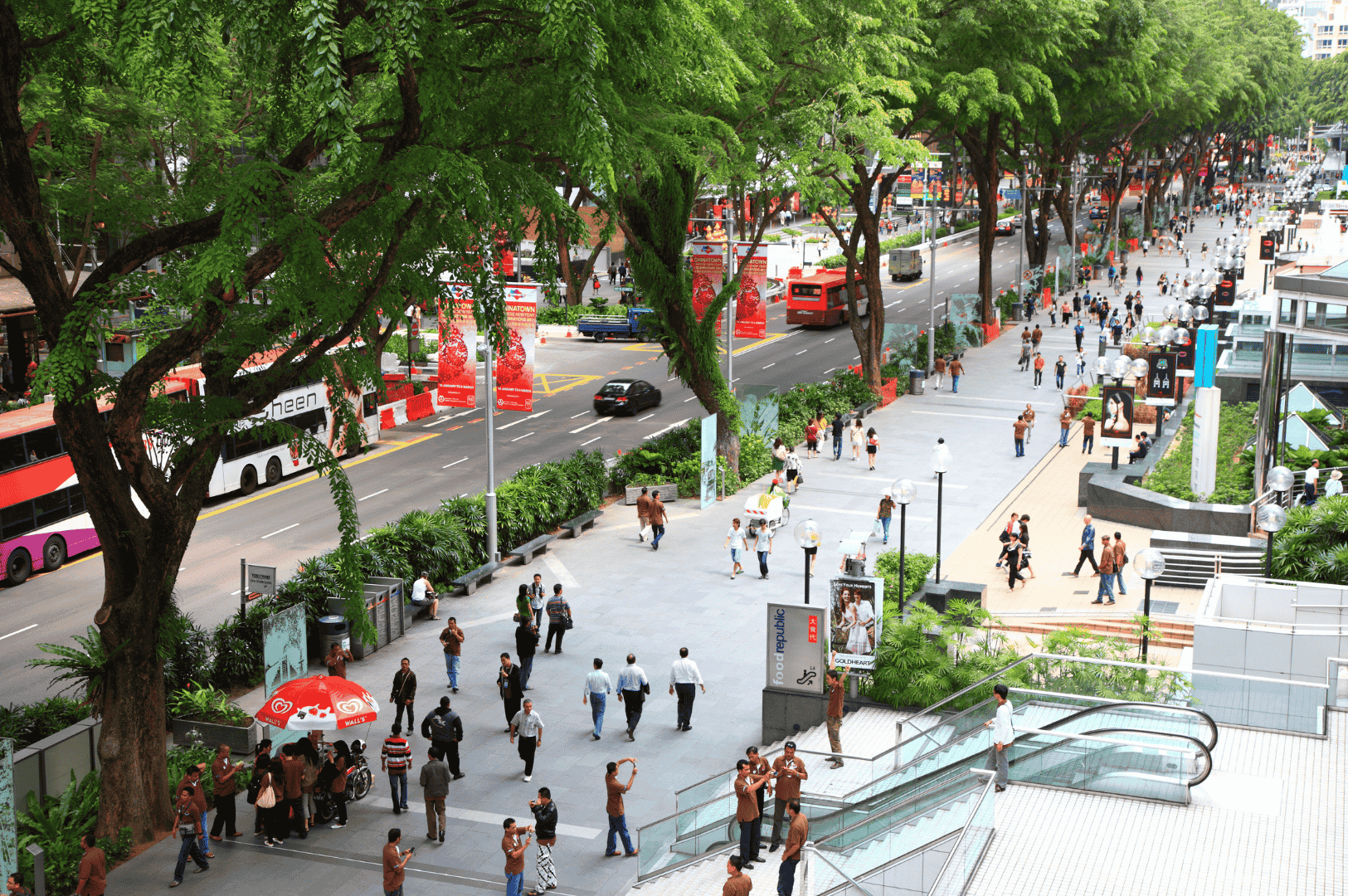 Wide pedestrian boulevard in a Singapore commercial district, lined with trees and red festive banners, as double-decker buses and crowds move through the organized, greenery-rich streetscape. This dynamic yet grounded scene reflects the city’s seamless blend of transit, retail, and public space beyond Orchard Road’s polished façade.