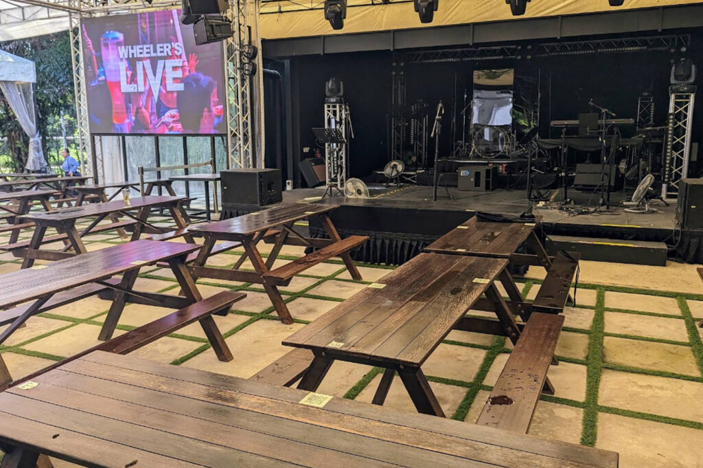 An outdoor performance area at Wheeler's Estate featuring long dark-wood picnic tables arranged on a concrete floor with a grass-grid pattern. In the background, a professional black stage is equipped with lights and a large LED screen that reads "WHEELER'S LIVE." The setting is airy and industrial-chic, located in a park-like environment.