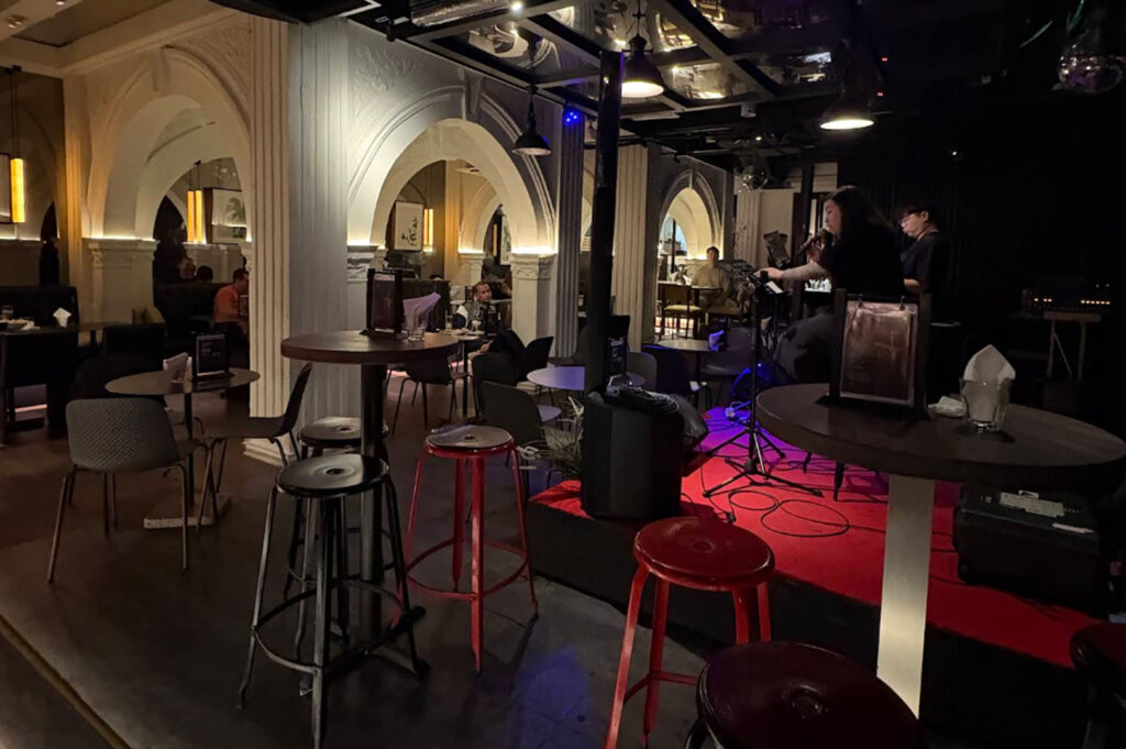 A cozy, dimly lit indoor music lounge featuring grand white architectural arches and pillars. In the foreground, several high-top wooden tables are paired with black and red metal stools. On a small, low-profile stage illuminated by a warm red glow, a female vocalist performs into a microphone. The background shows more intimate seating areas under vaulted ceilings, creating a sophisticated yet relaxed atmosphere.