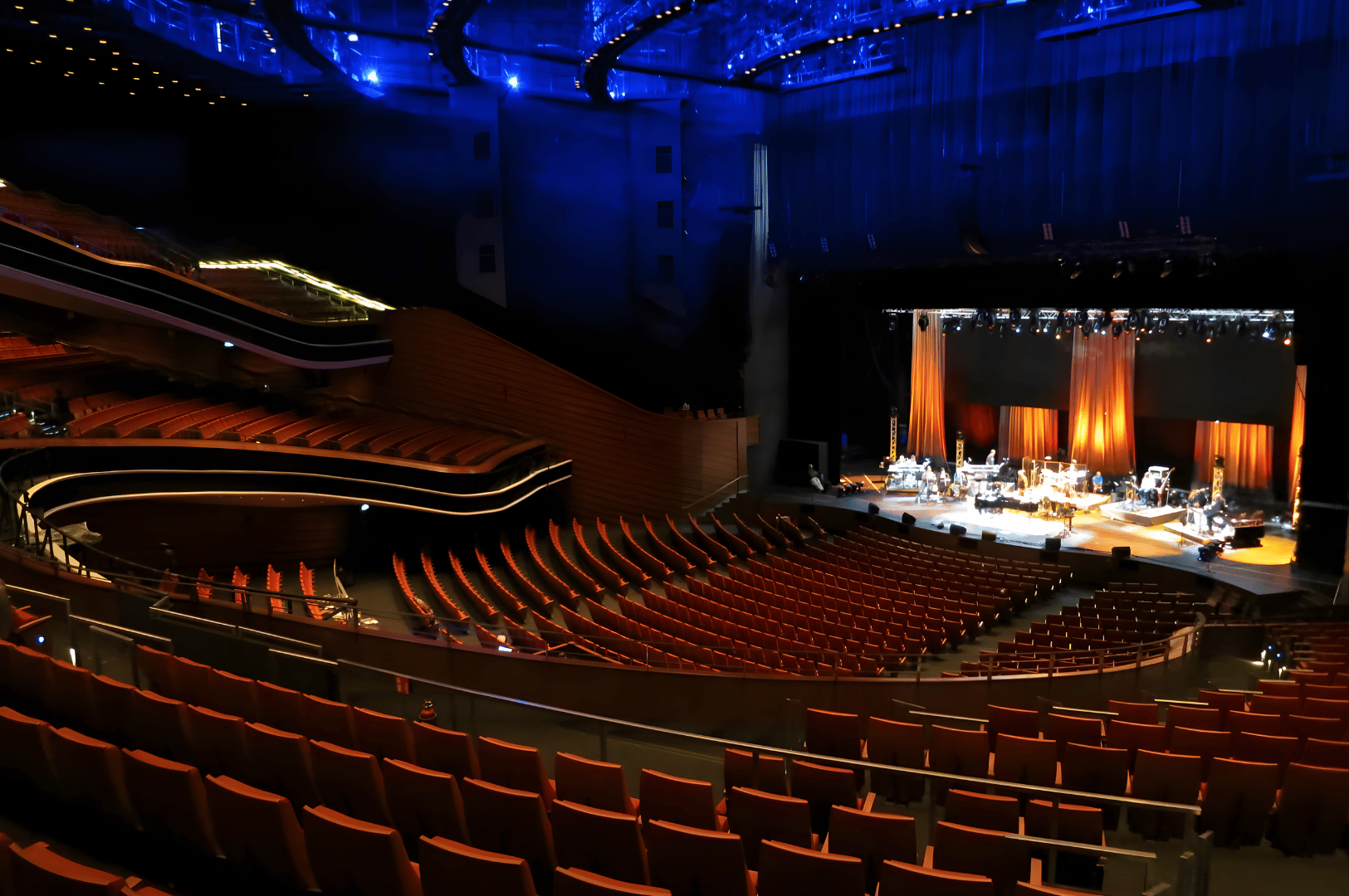 Modern Singapore concert hall with tiered seating and sleek architectural design, audience facing a brightly lit stage where musicians perform under blue ambient lighting, reflecting the scale and sophistication of the city’s live music venues.