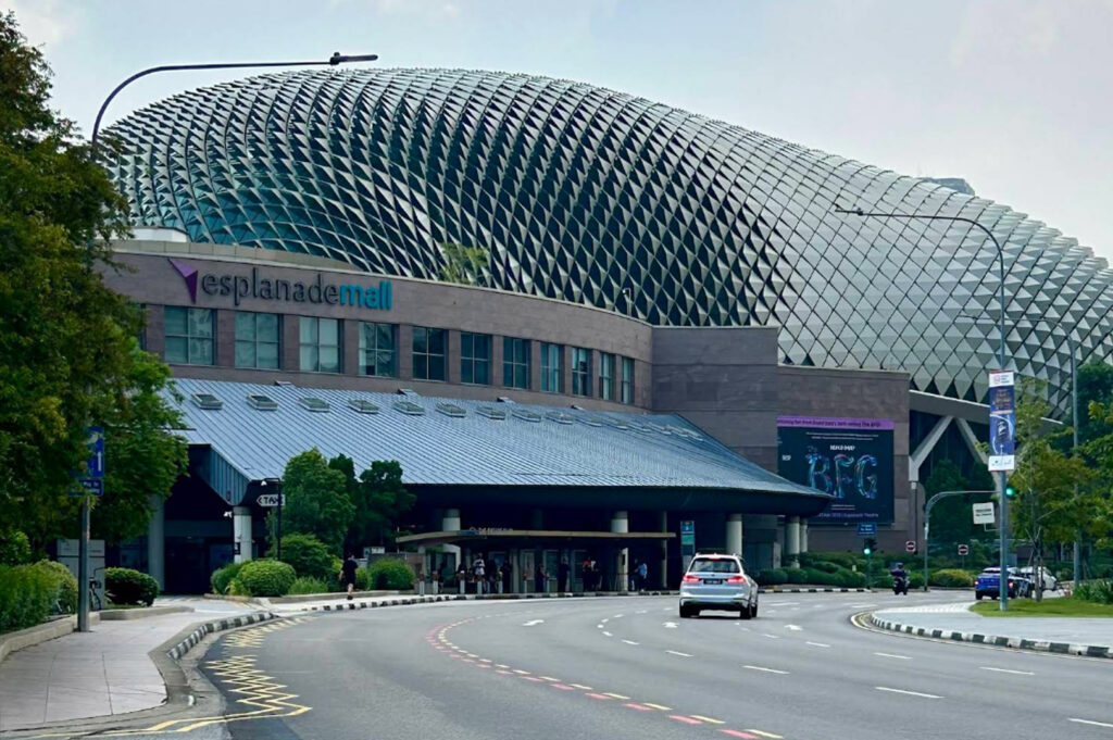 A daytime exterior shot of the Esplanade Mall in Singapore. The building features a prominent, modern "durian-like" architectural dome covered in thousands of triangular aluminum sunshades. A curved road with a silver SUV leads toward the entrance. Lush green trees line the sidewalk, and a purple digital billboard is visible on the side of the building.
