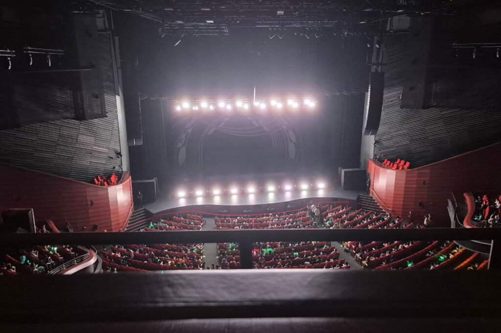A high-angle view from the balcony of a large, modern concert hall. Rows of red theater seats are filled with an audience facing a brightly lit stage. The stage features a series of powerful spotlights creating a hazy glow. The walls of the theater are tiered with dark, textured wood paneling and curved red balconies, emphasizing the scale of the venue.