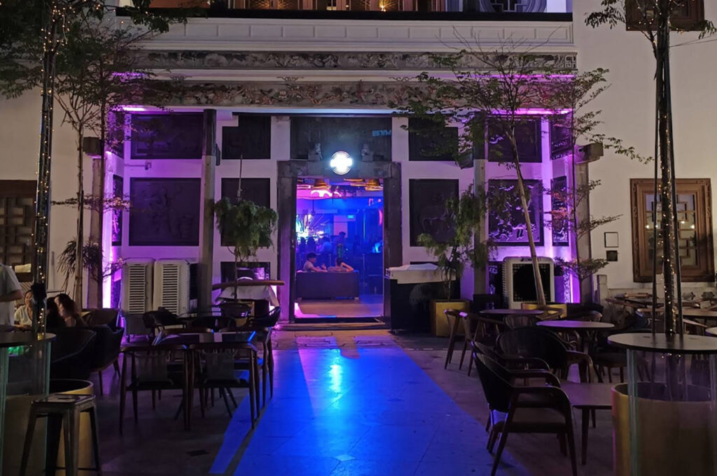 An outdoor courtyard at night, illuminated by vibrant purple and blue neon lights. The entrance to a building is framed by traditional-style stone carvings and pillars. In the foreground, several dark round tables and chairs are set up for patrons. Fairy lights are wrapped around thin trees, adding to the energetic, nightlife ambiance.