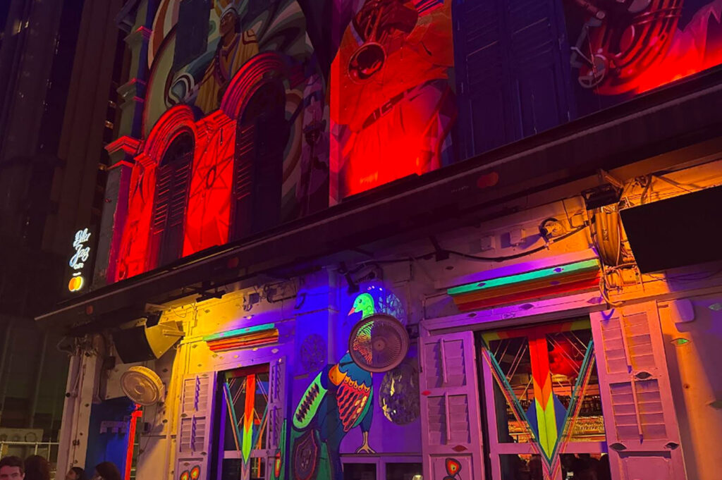 A close-up, low-angle shot of the colorful exterior of Blu Jaz Cafe. The building is covered in vibrant, multi-colored murals depicting a peacock and jazz musicians. The scene is saturated with intense red, blue, and yellow light from overhead street lamps and neon signs, highlighting the eclectic and artistic character of the Haji Lane area.