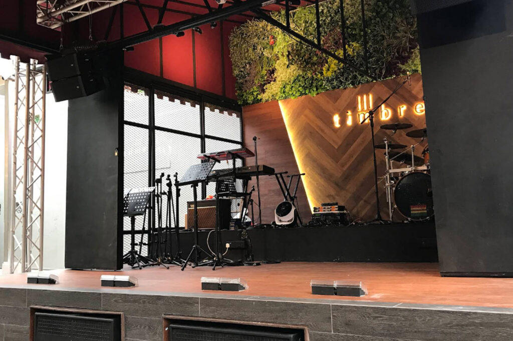 A clean, well-lit view of a live music stage at Timbre. The stage features a light-colored wooden floor and a backdrop with a "V" shaped wooden slat design and a wall of green foliage. A neon "timbre" sign glows warmly on the back wall. Musical equipment, including a drum kit, keyboards, and microphone stands, is neatly arranged and ready for a performance.