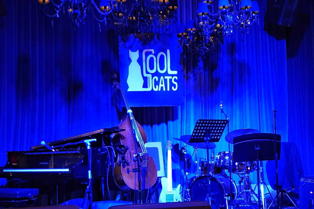 An intimate stage at Cool Cats jazz club, bathed in deep blue light. A large "COOL CATS" logo featuring a cat silhouette hangs at the center. The stage is packed with professional instruments, including a grand piano, a double bass, a drum set, and an electric guitar. Ornate crystal chandeliers hang from the ceiling, casting a classic, high-end jazz lounge vibe.