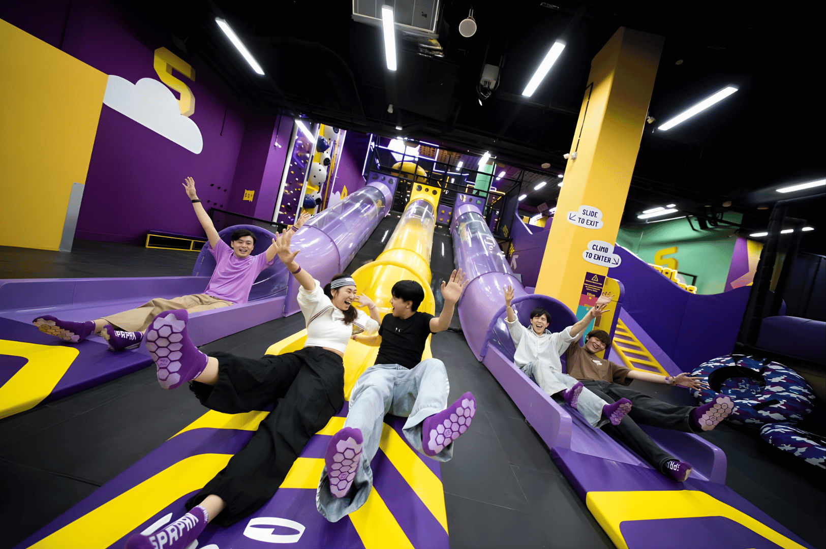 Five individuals gleefully slide down vibrant purple and yellow indoor slides, arms raised in excitement amid a lively, modern play facility—an energetic snapshot of Singapore’s growing landscape of indoor family attractions that go beyond the blockbuster thrills of Universal Studios.