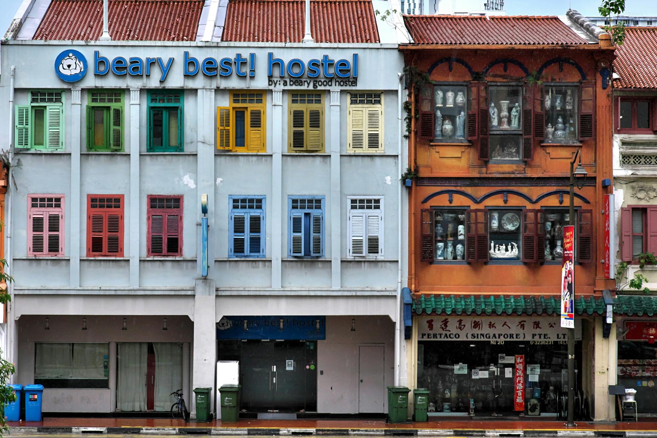 This vibrant street scene features a row of heritage shophouses, contrasting a light blue building adorned with a grid of multicolored shutters against a neighboring structure with a warm, ornate orange facade.