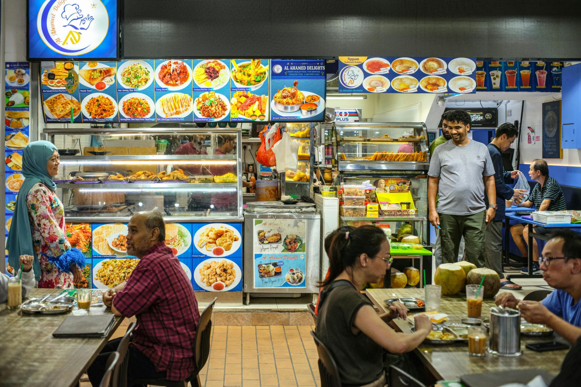 Patrons are seated at tables enjoying meals and drinks in a lively, brightly lit indoor food court. Behind them, a stall named "Al Ahamed Delights" displays a colorful menu of dishes above a glass counter filled with food, where a woman in a floral headscarf stands nearby.