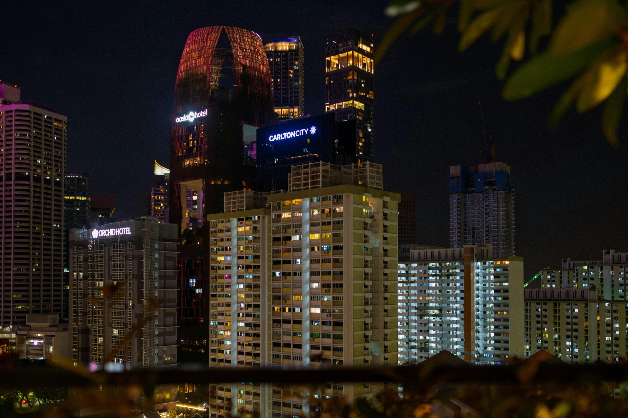 This night cityscape captures a dense urban skyline where the illuminated "Oasia Hotel" and "Carlton City" signs stand out atop modern skyscrapers. In the foreground, the warm glow of lights from numerous windows in tall residential apartment blocks creates a striking contrast against the dark sky.