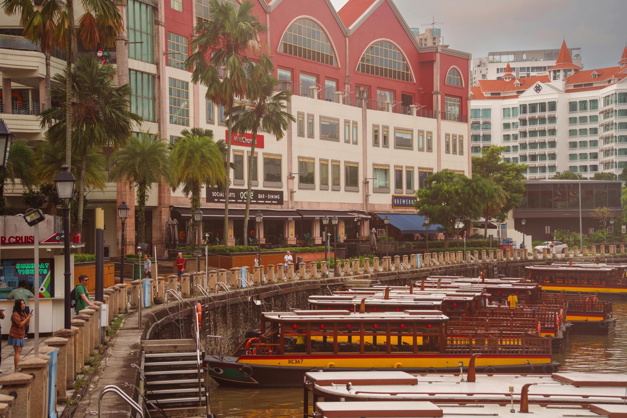 A fleet of traditional wooden tour boats sits docked along a curved riverbank promenade lined with palm trees and lampposts. Behind the waterfront walkway, large multi-story buildings with distinctive red roofs and arched windows house various dining establishments like "Social Place" and "Brewerkz".
