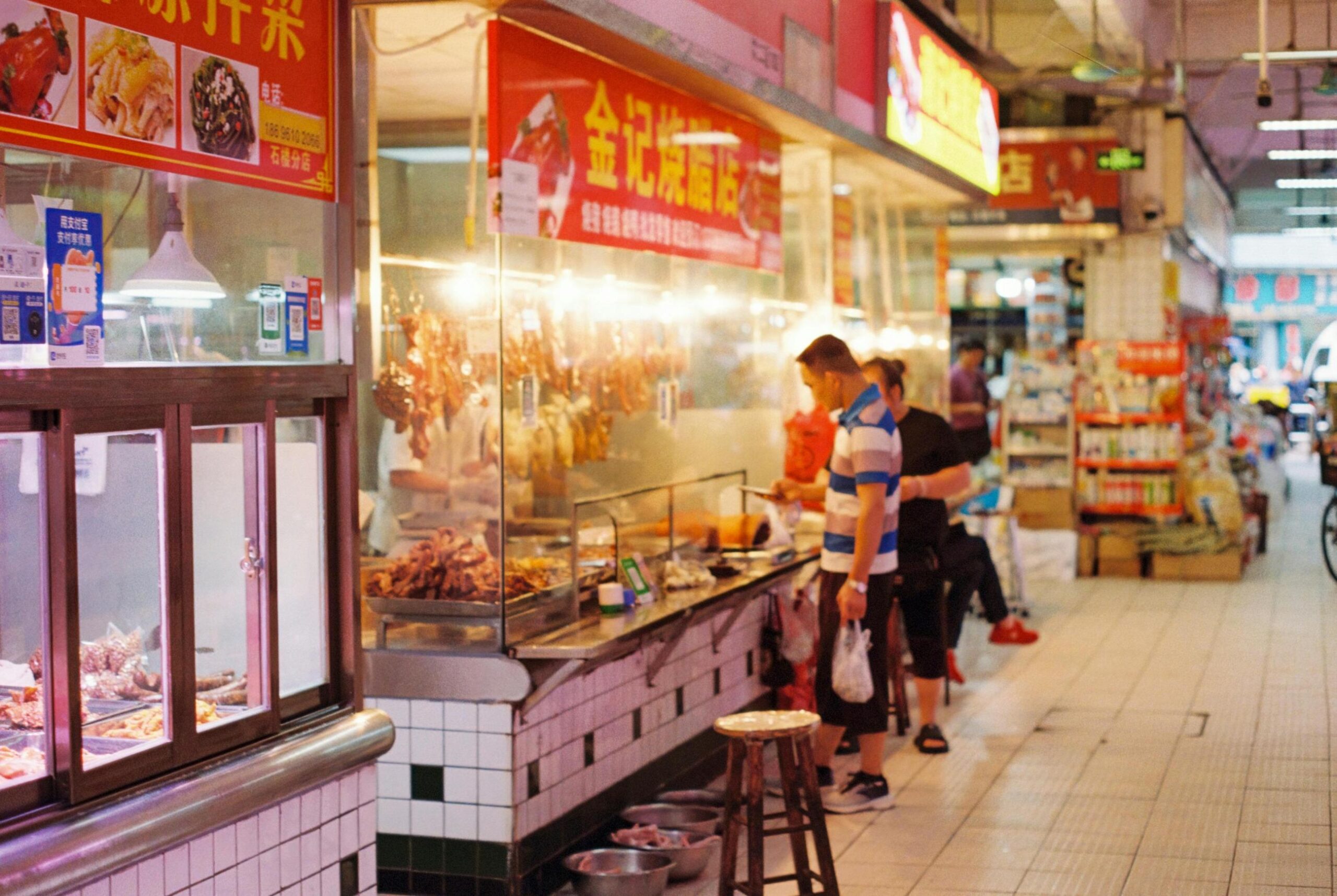 A man in a striped shirt waits at a bright, indoor Chinese food stall featuring hanging roasted meats and red signage.