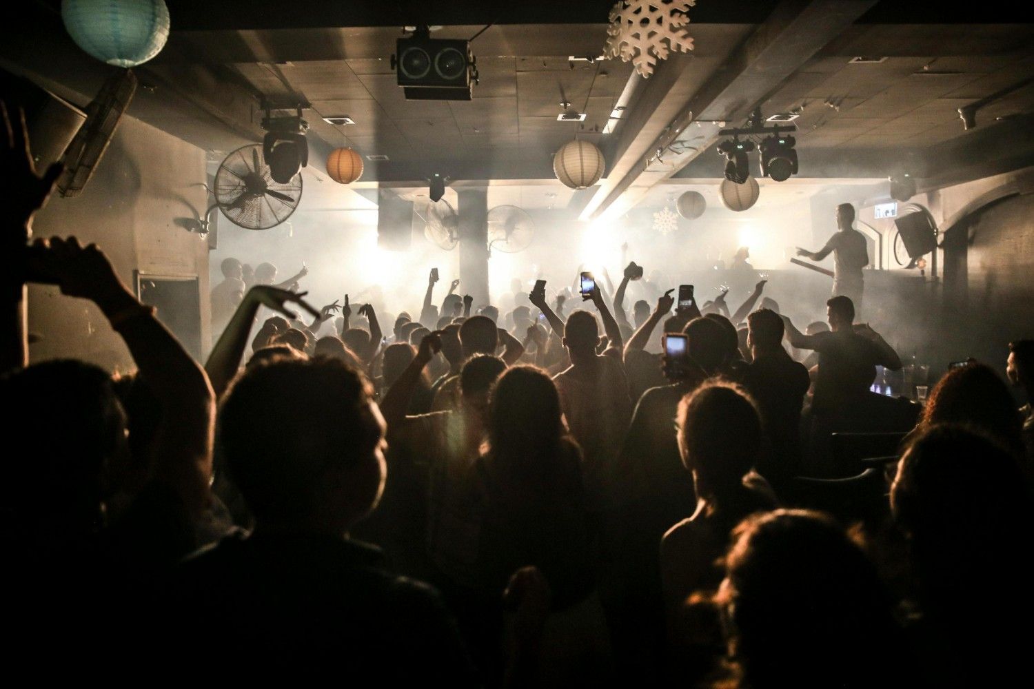  A large crowd of people is gathered in a dark, hazy nightclub, with many individuals raising their hands and holding up smartphones to capture the event. Bright spotlights from the stage illuminate the silhouetted audience and the various paper lanterns and decorations hanging from the ceiling.