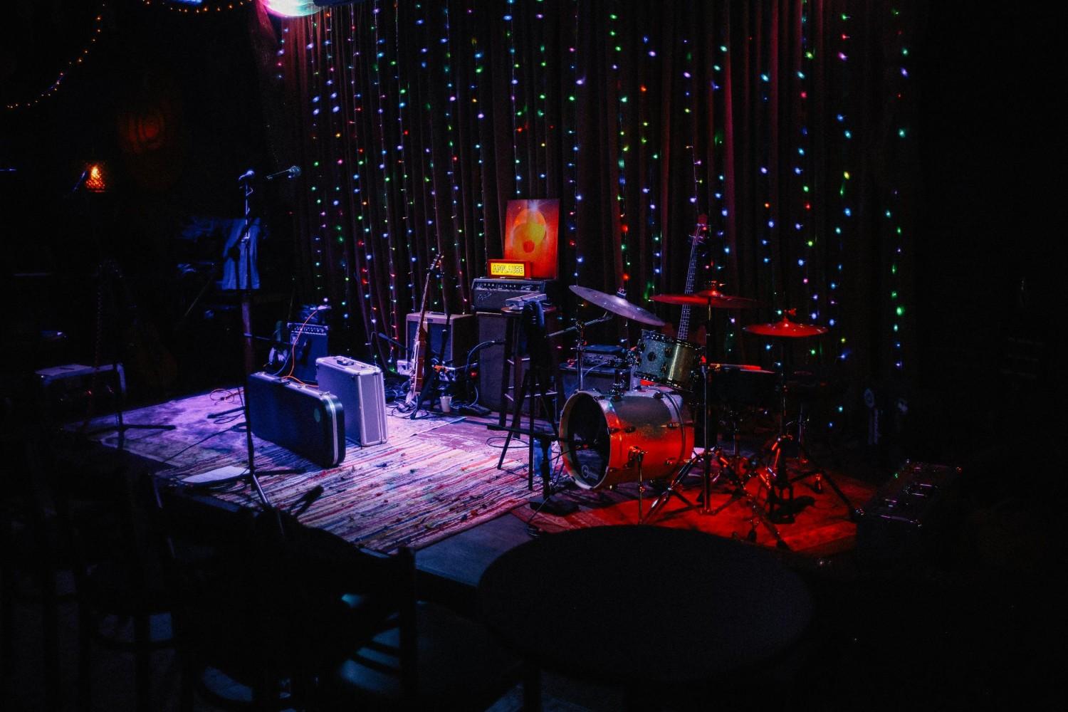  A small, intimate stage is set up with a drum kit, guitars, and equipment cases resting on a colorful rug. Behind the instruments, a dark curtain is draped with vertical strings of multi-colored fairy lights, creating a festive backdrop for a performance.