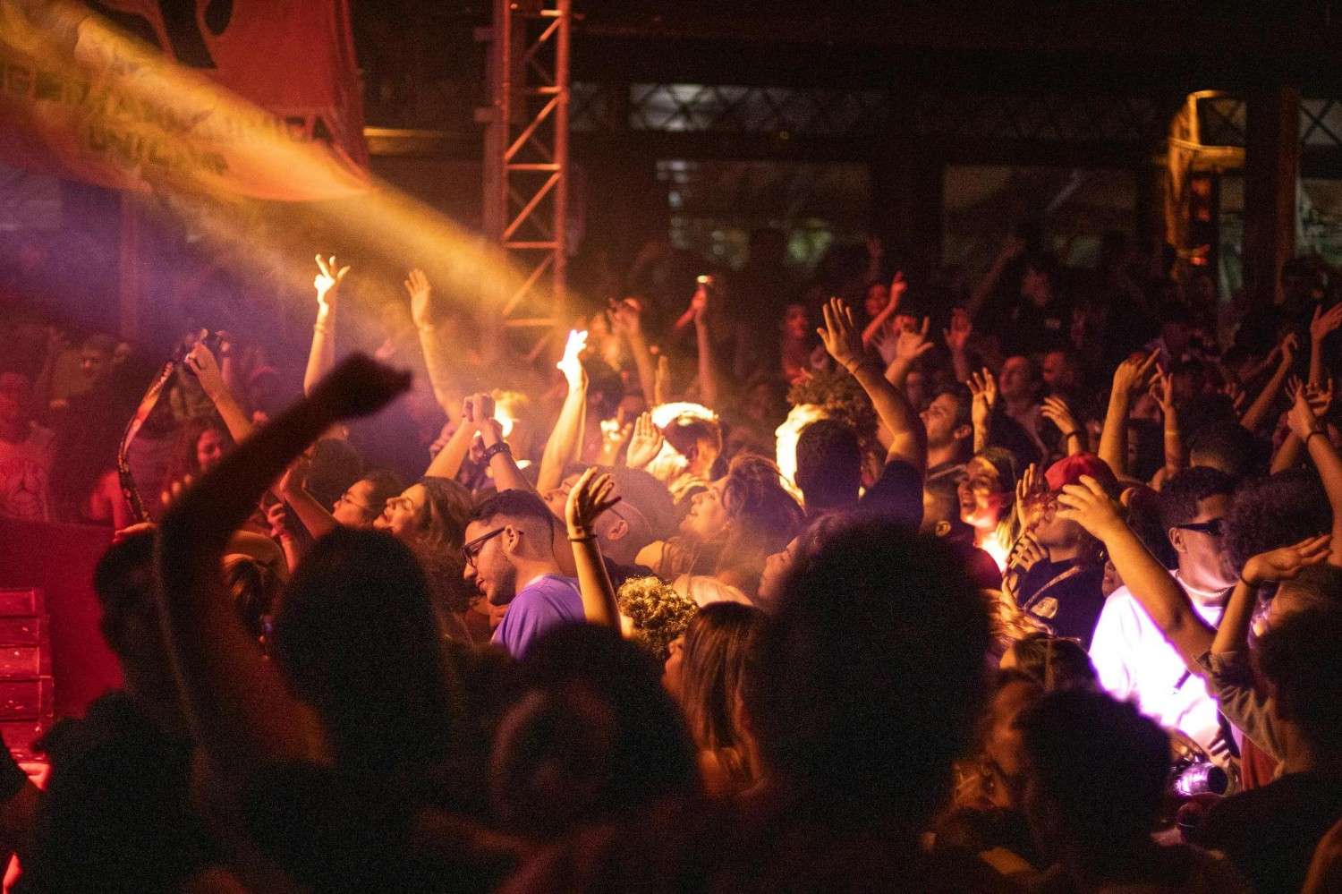  A vibrant crowd of people is gathered in a dimly lit venue, with many individuals raising their hands and cheering in the warm glow of stage lights. A bright beam of light cuts through the hazy atmosphere, illuminating the faces of the audience members as they enjoy the live event.