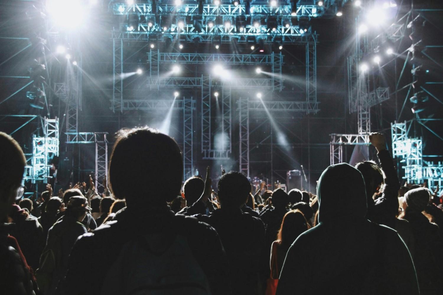A large audience is seen from behind, gazing toward a massive stage structure equipped with intricate metal scaffolding and bright white spotlights. The crowd is silhouetted against the intense beams of light, creating a sense of anticipation and scale at a major outdoor or arena concert.
