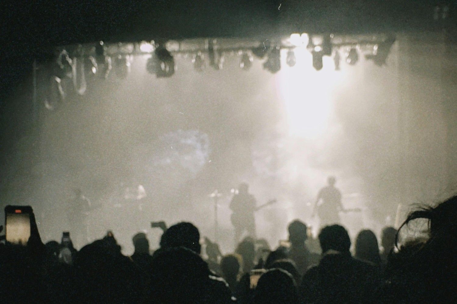  A band performs on a hazy stage in front of a large, silhouetted audience, with musicians visible as dark figures against a bright white light source. The image has a grainy, film-like quality, capturing the energy of the crowd as several people hold up smartphones to record the live performance.