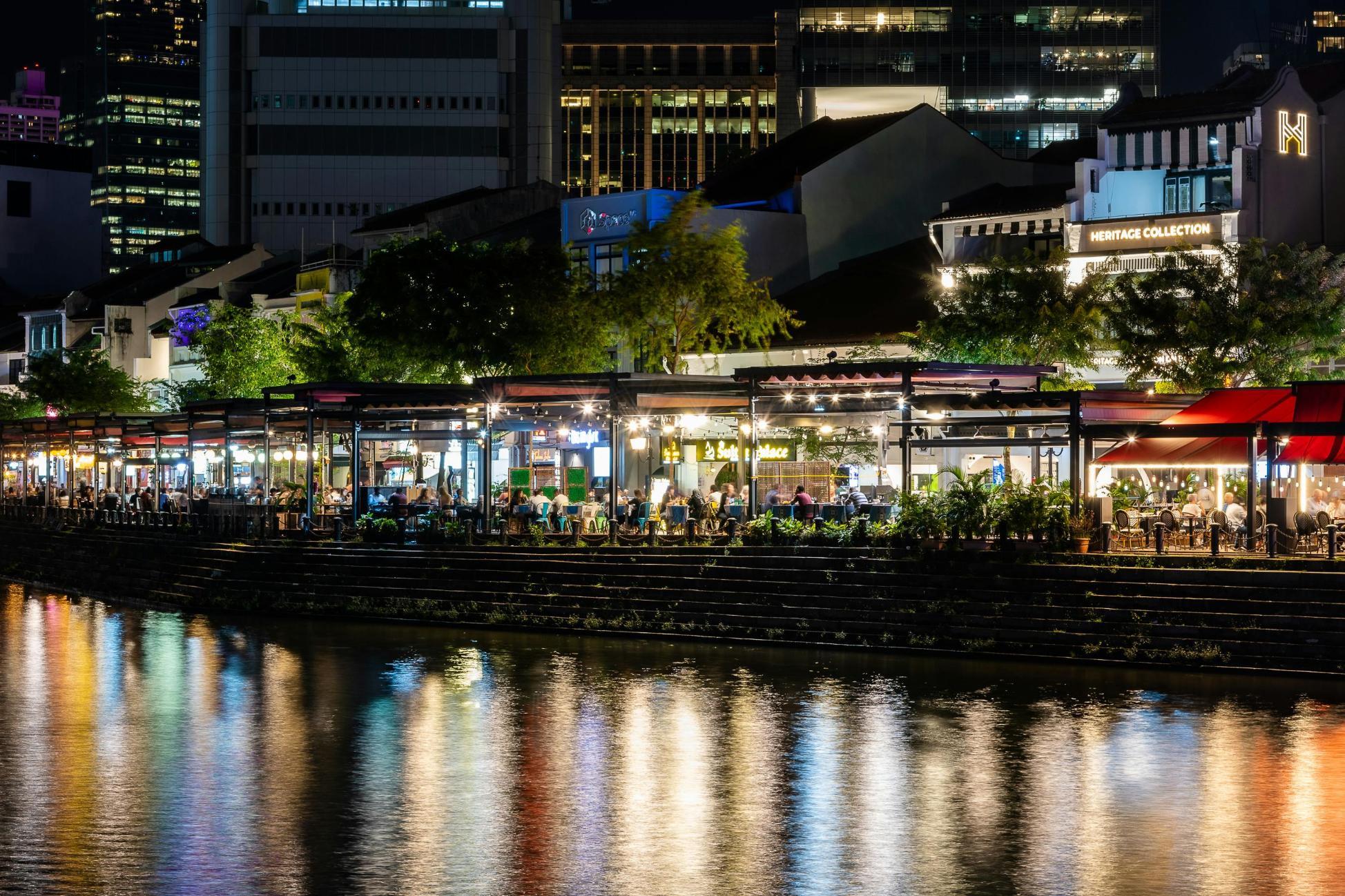  This is a captivating, long-exposure night photograph showcasing a row of brightly lit riverside restaurants and bars, likely in a city setting.