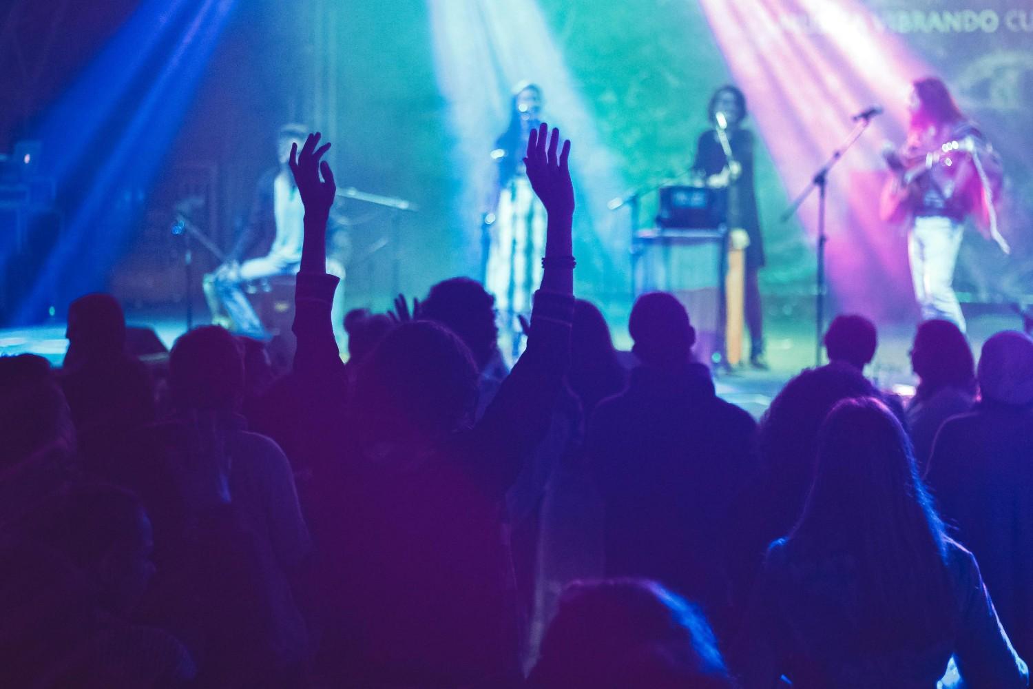 Silhouetted audience members watch a live performance on a stage illuminated by dramatic beams of blue and purple light. A central figure in the crowd raises their hands high, capturing the energetic atmosphere of the concert while musicians perform in the hazy background.