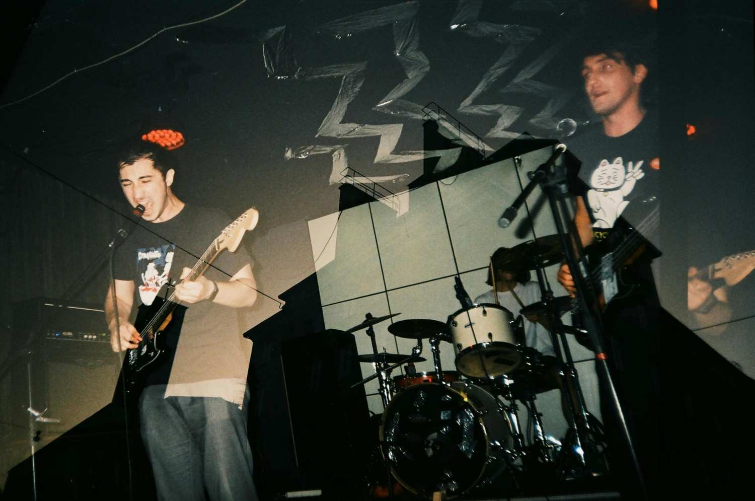 Captured with a creative multi-exposure effect, a rock band performs live, featuring a guitarist singing into a microphone on the left and a bassist wearing a graphic tee on the right. The gritty, low-light composition includes a drummer in the background and jagged white zig-zag patterns on the ceiling, emphasizing the raw energy of the performance.