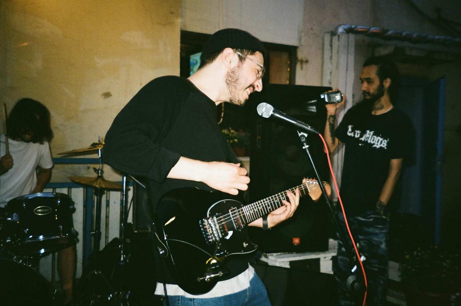 A smiling guitarist wearing a black beanie and glasses plays a black electric guitar, leaning towards a microphone during an intimate performance. Behind him, a drummer focuses on their kit while another individual stands to the side, capturing the show with a handheld camcorder.
