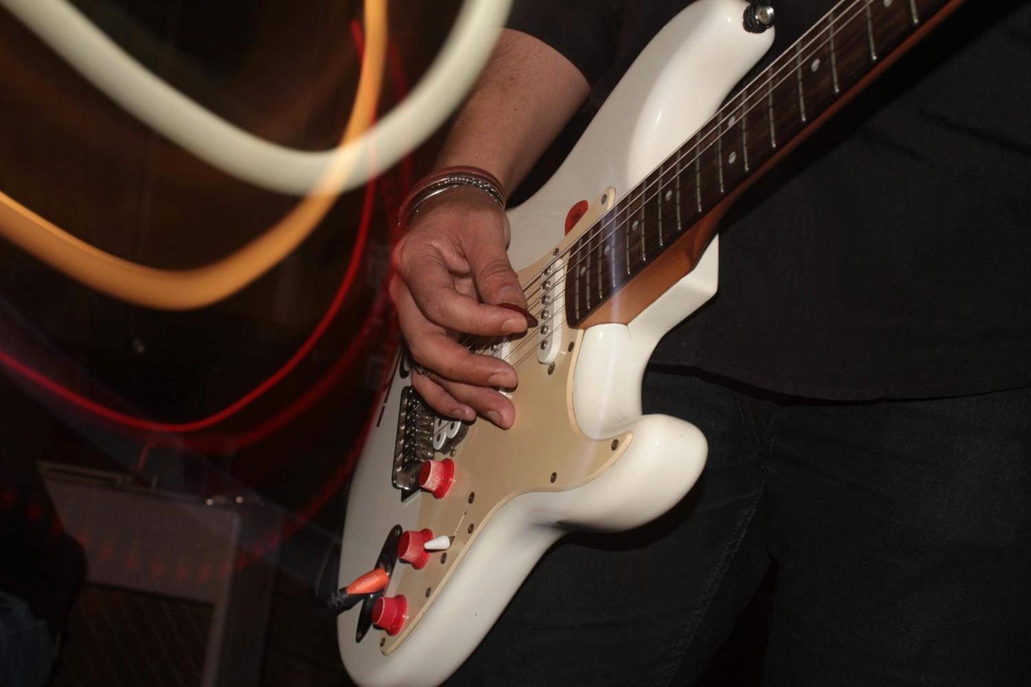 A close-up shot focuses on a musician's hands playing a white electric guitar characterized by distinctive red control knobs. Swirling trails of yellow and red light curve across the foreground, adding a dynamic, motion-blurred effect to the musical performance.