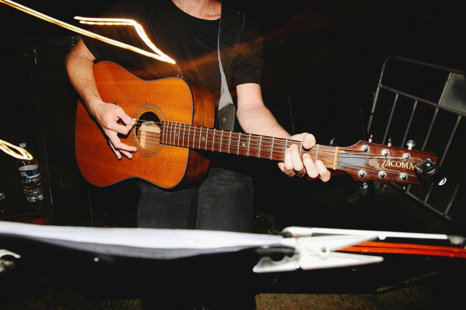 A musician wearing a black t-shirt strums a wooden acoustic guitar with the "Tacoma" logo clearly visible on the headstock. Bright, motion-blurred streaks of yellow light cut across the dark scene, adding dynamic energy to the performance above a music stand in the foreground.